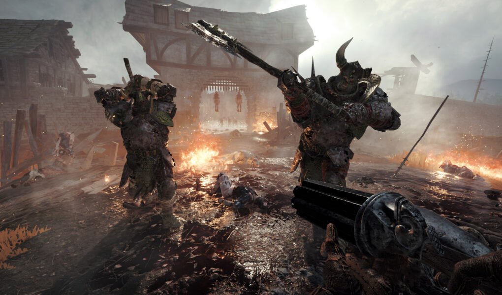 Two armoured Chaos Warriors advance through a burning village while the player aims a gun in Vermintide 2.