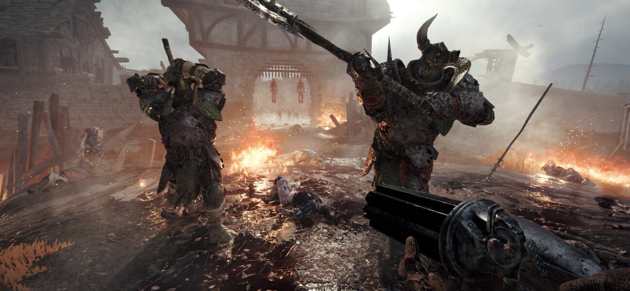 Two armoured Chaos Warriors advance through a burning village while the player aims a gun in Vermintide 2.